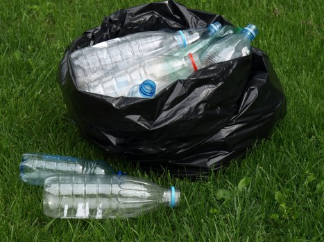 A partially filled black rubbish bag sits on a grassy area, containing several clear plastic water bottles with blue caps and one red-marked bottle, some of which are spilling out onto the ground. The plastic bottles are of varying sizes and are mostly compressed, showcasing their transparent, smooth, and slightly reflective surfaces. The scene appears to be outdoors, possibly on a residential driveway or garden space, with vibrant green grass surrounding the waste. The bottles and bag are positioned centrally in the image, with natural sunlight highlighting the textures and transparency of the plastic. This visual is representative of rubbish collection or waste removal services, such as those offered by House Clearance Broxbourne, which handle household rubbish including plastic bottles and general waste disposal in the Broxbourne area.
