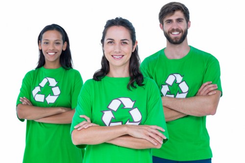 A group of three modern-looking individuals, two women and one man, standing outdoors with arms crossed, wearing bright green T-shirts featuring a large white recycling symbol. The woman in the middle has dark hair pulled back and is smiling directly at the camera. To her left, another woman with long dark hair is also smiling, while the man on the right has short dark hair, a beard, and is smiling as well. The background appears to be an outdoor setting, possibly in a residential or community area, with no distinct details visible. Their presence suggests they are part of an environmental or waste management initiative, aligning with rubbish removal services offered by groups like House Clearance Broxbourne, based near Broxbourne in Hertfordshire.