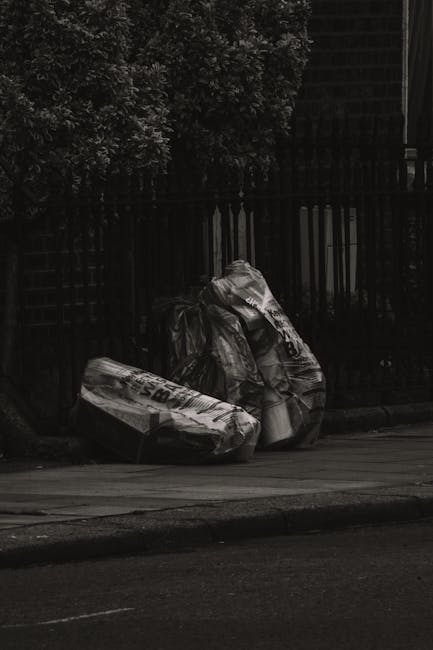 Two large black plastic rubbish bags, filled and tied at the top, are placed on a street pavement in front of a dark wooden fence and leafy bushes. The bags appear to contain household waste or garden