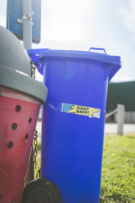 Two large black plastic rubbish bags, filled and tied at the top, are placed on a street pavement in front of a dark wooden fence and leafy bushes. The bags appear to contain household waste or garden