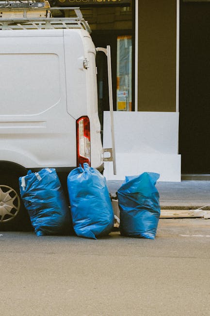 A collection of discarded rubbish piled on the ground beside a stone wall and metal fence, with a background featuring a parked vehicle and a blue sky with light clouds. The rubbish includes black pla
