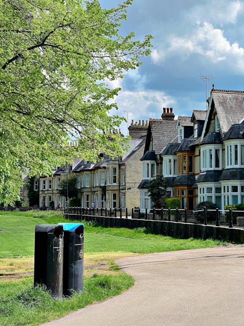 A wide view of a suburban residential area featuring a row of traditional Victorian-style terraced houses with sash windows, bay windows, and slate roofs. In the foreground, a paved pathway runs paral