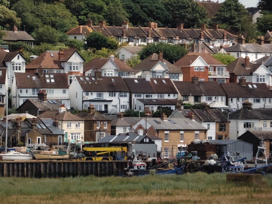 A wide view of a suburban residential area featuring a row of traditional Victorian-style terraced houses with sash windows, bay windows, and slate roofs. In the foreground, a paved pathway runs paral
